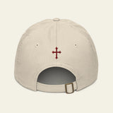 A light beige baseball cap shown from the back, featuring a small embroidered dark red cross on the center back and an adjustable strap with a metal buckle.